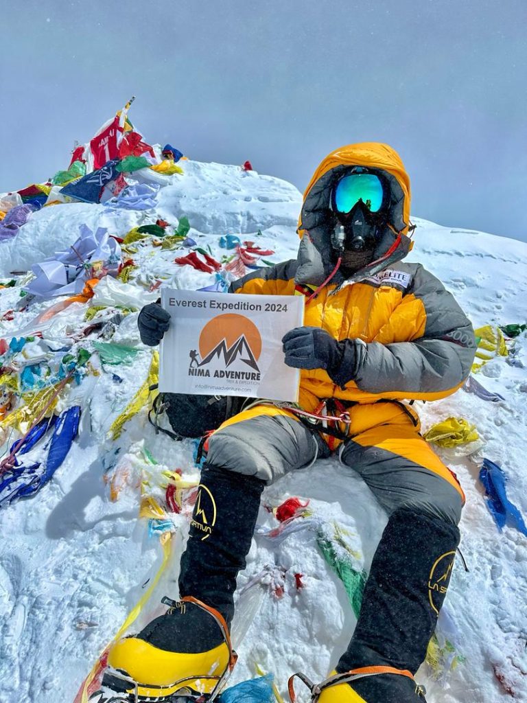 Climber on the summit of Everest holding a Nima Adventure banner among prayer flags