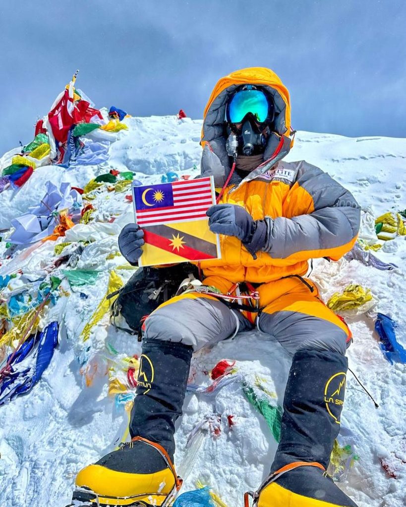 Climber on the summit of Mount Everest (8,849 m) holding the Malaysian and Sarawak flags, surrounded by prayer flags