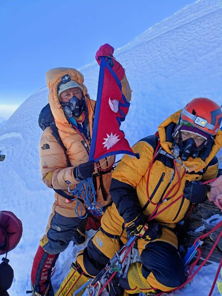 Two climbers in down suits on a snowy summit with the flag of Nepal