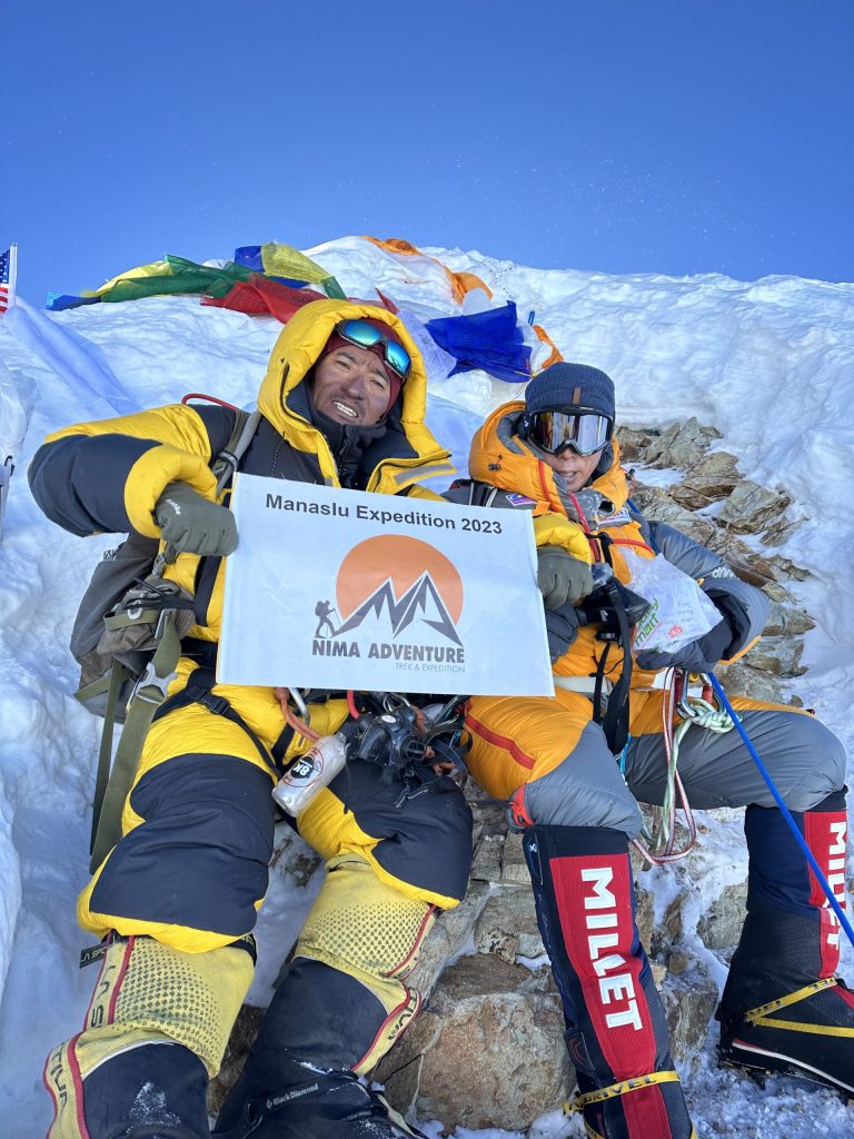 Climber on a snowy summit holding a Nima Adventure banner among prayer flags