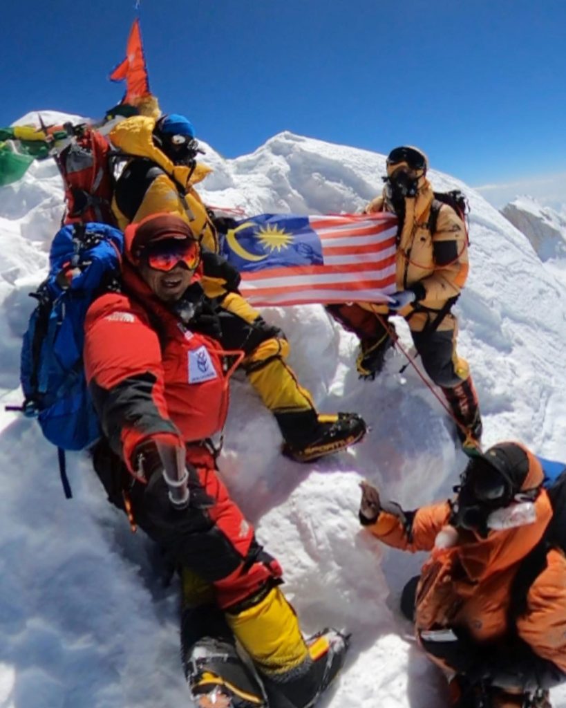 Climbers on the snowy true summit of Manaslu (8,163 m) holding the Malaysian flag under a blue sky