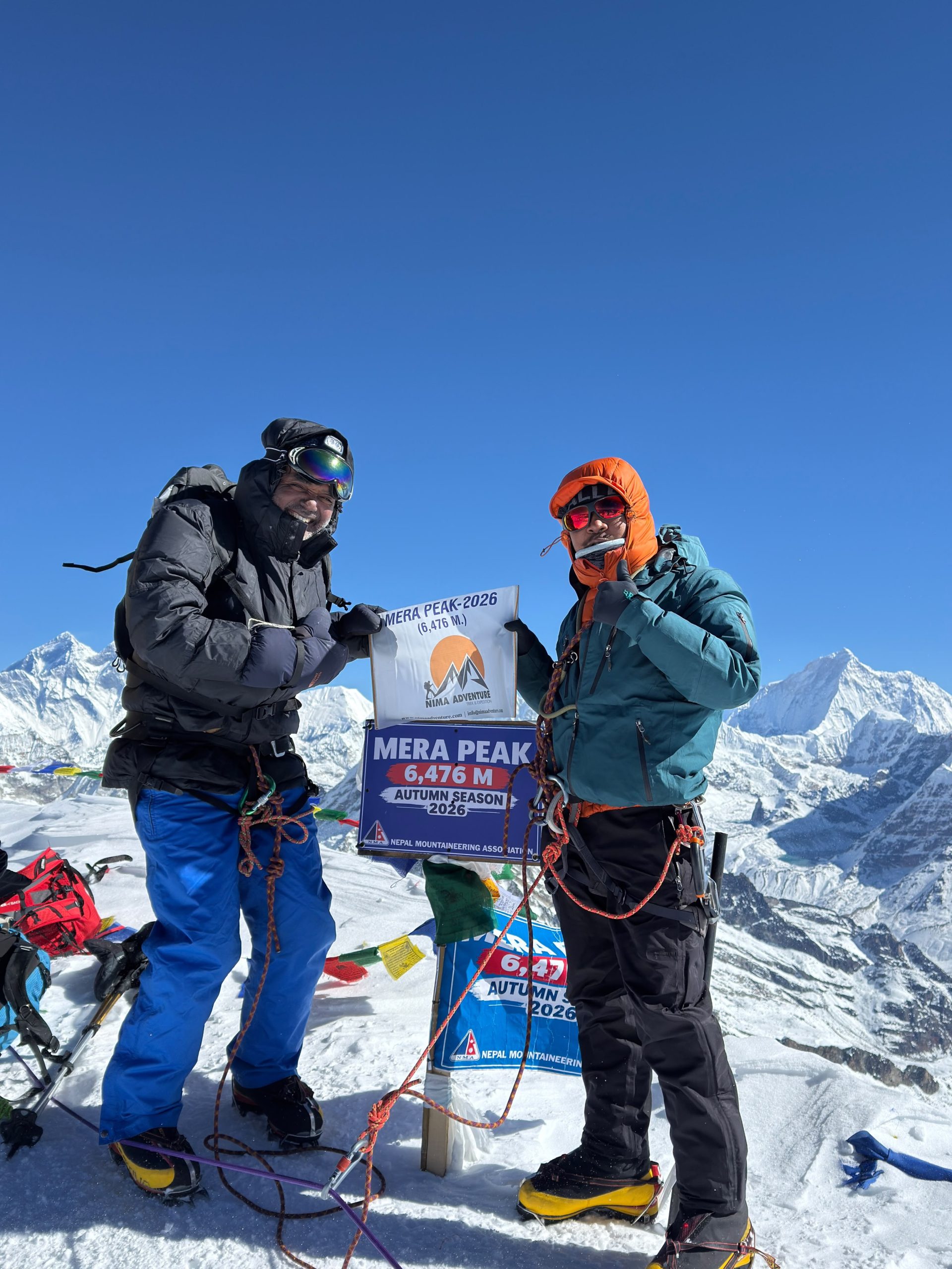 Two climbers in down jackets beside the Mera Peak 6,476 m summit sign, with Himalayan peaks behind