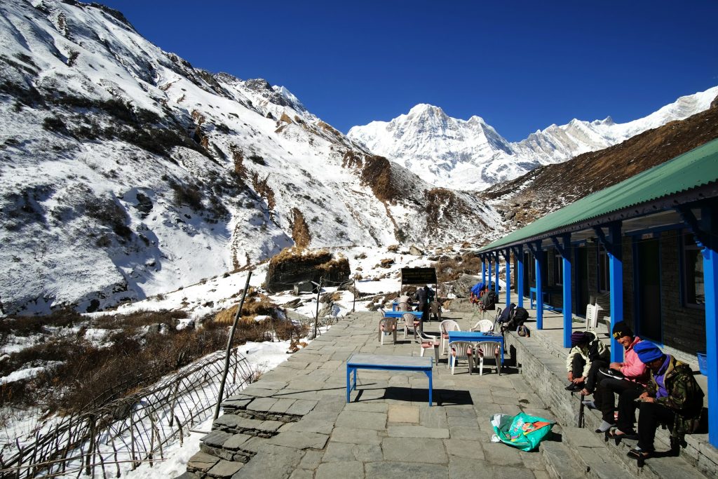 Trekkers resting on a stone terrace outside a blue-pillared lodge in a snowy valley in the Annapurna Sanctuary