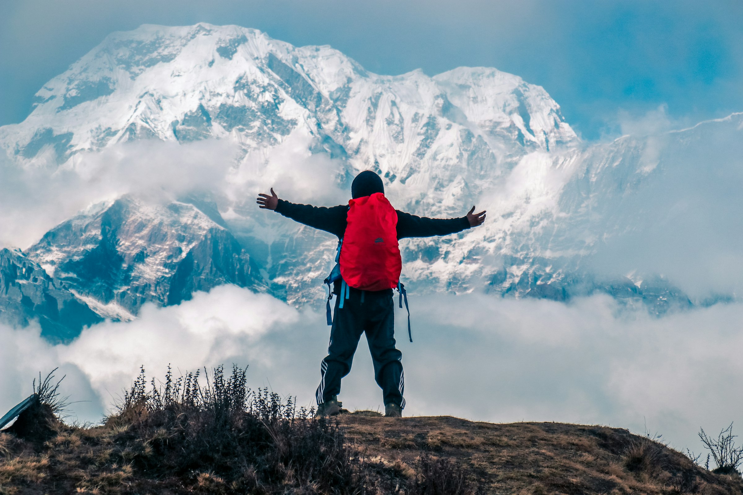 Trekker with a red backpack raising both arms towards a huge snowy massif above the clouds