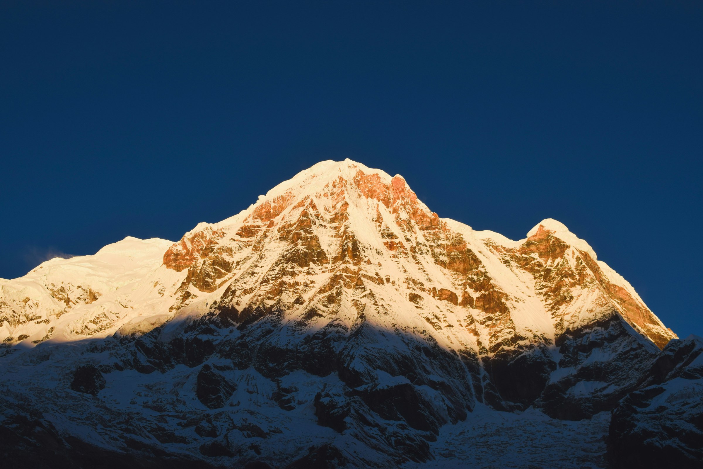 Snowy mountain face lit golden by early sun against a deep blue sky in the Annapurna Sanctuary