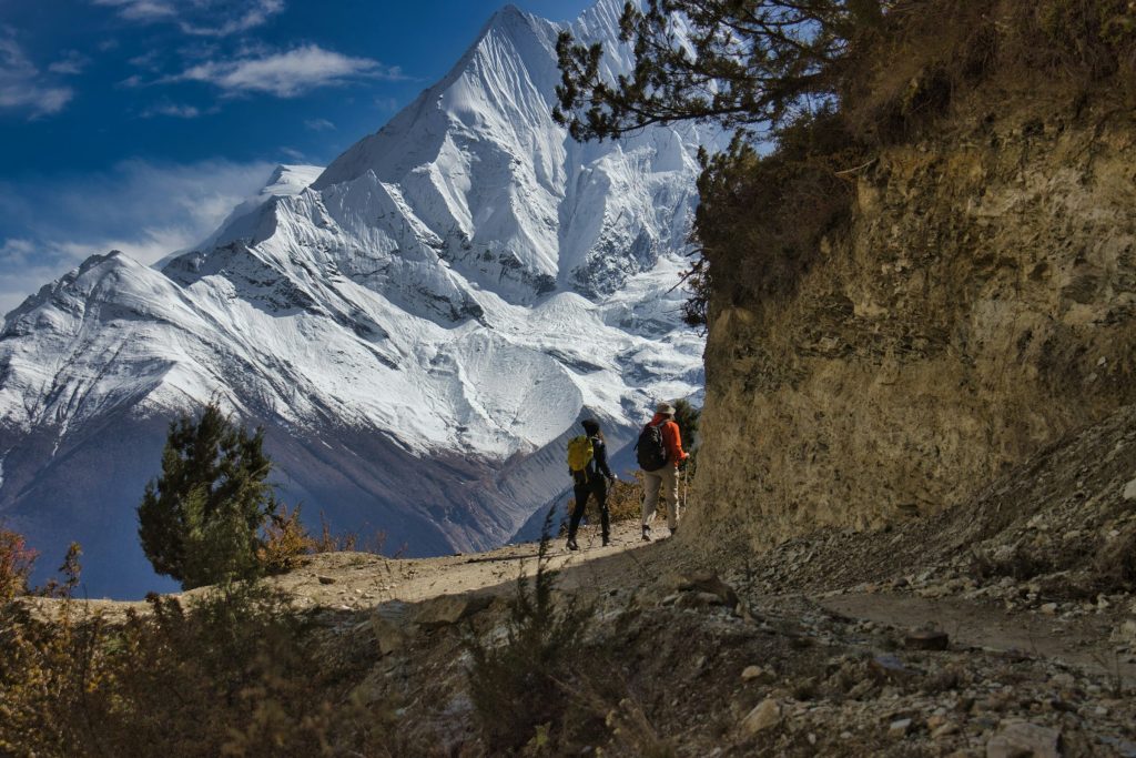 Two trekkers on a cliffside trail with a huge snowy peak towering behind in the Annapurna region