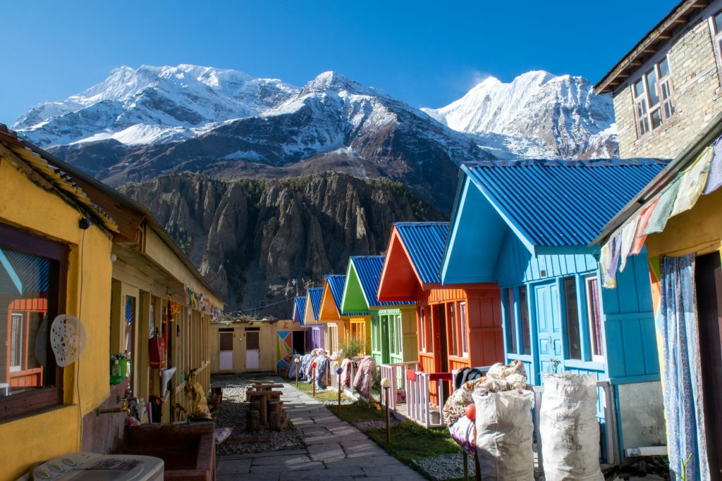 Row of brightly painted lodge rooms beside a stone path below snowy peaks in the Annapurna region