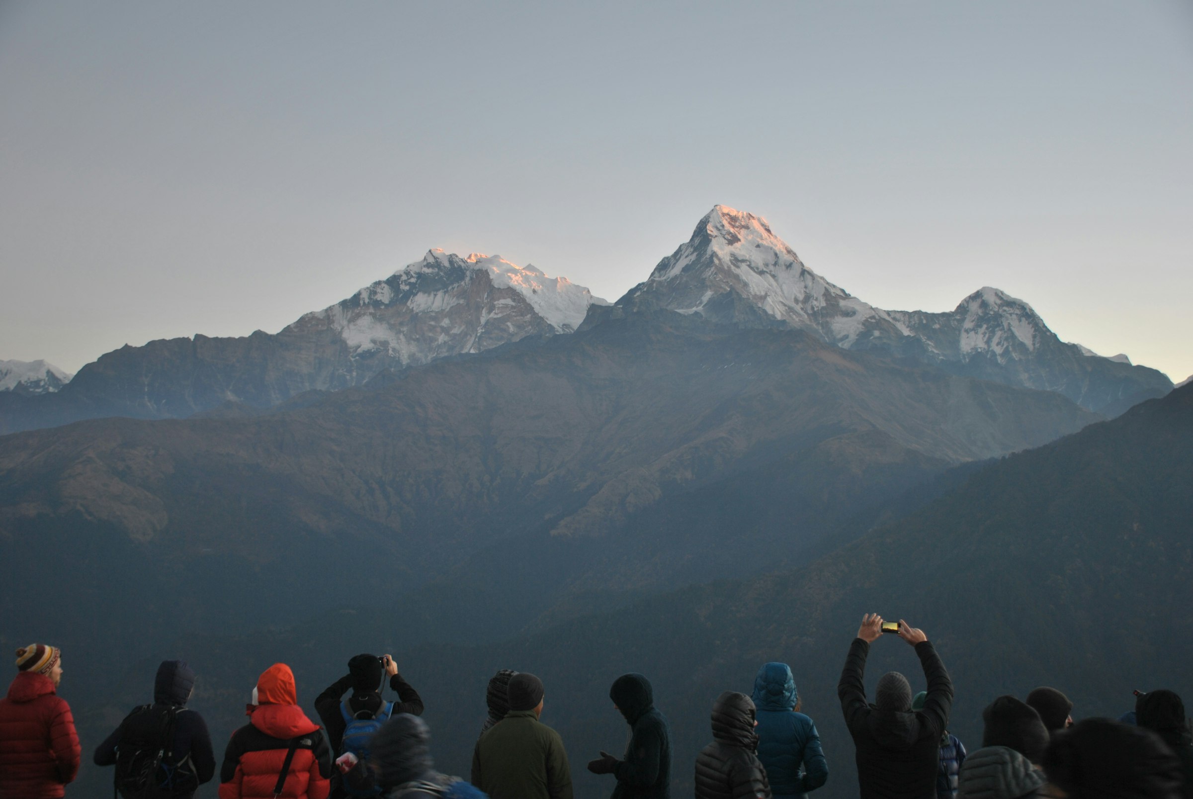Trekkers silhouetted at dawn photographing sunrise over snowy Annapurna peaks from a hilltop viewpoint