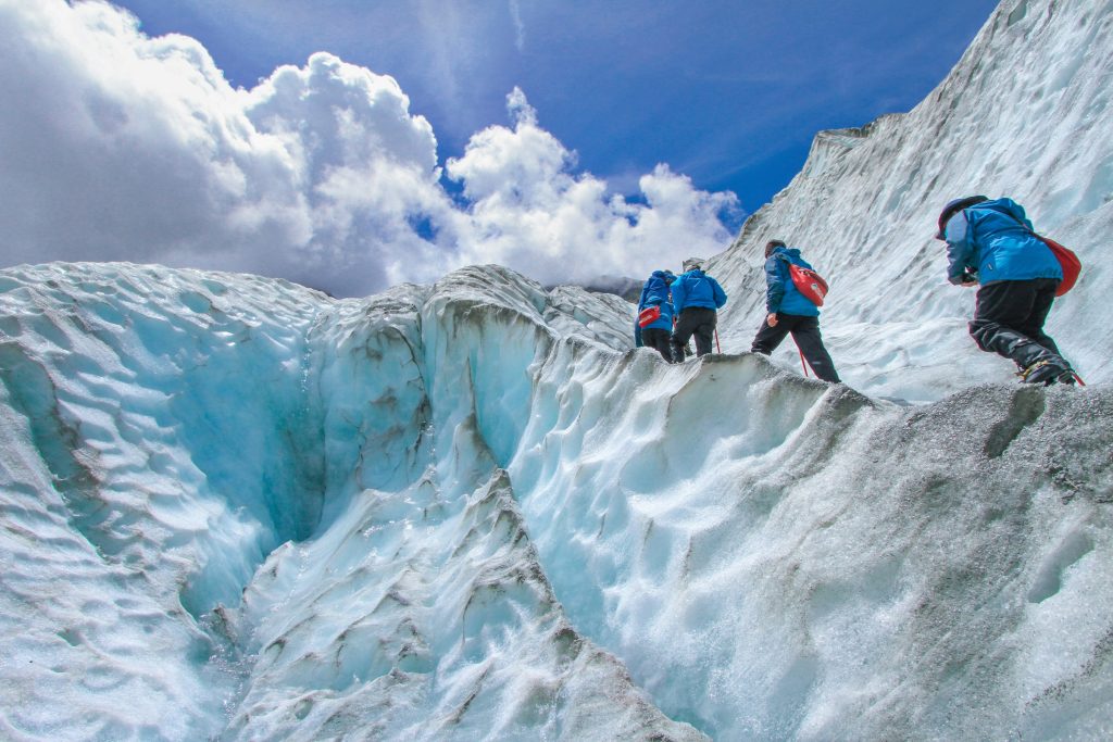 Four climbers in blue jackets scrambling across ridged blue glacier ice under a cloudy sky