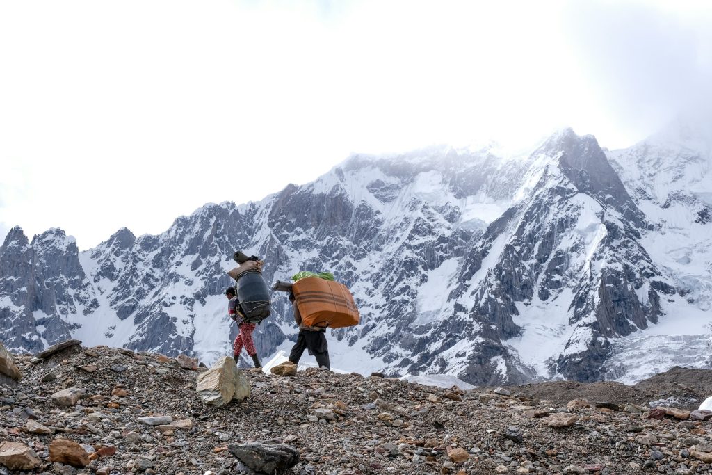 Two porters carrying large loads across rocky moraine below snowy mountains in the Karakoram