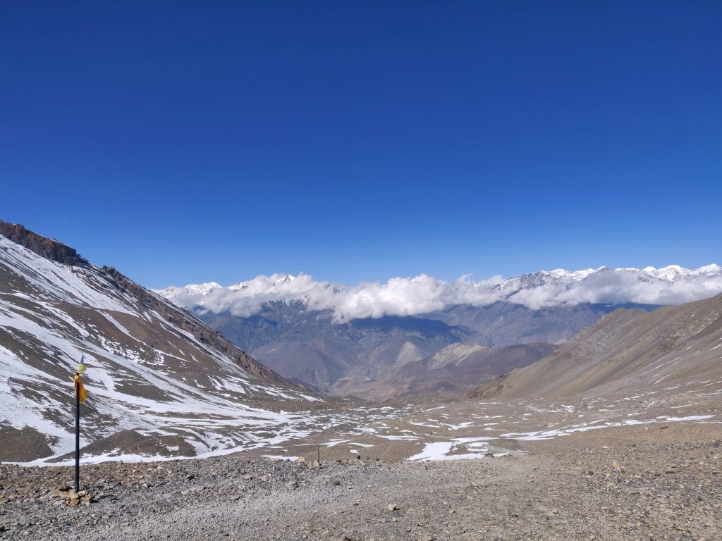 Wide snow-patched valley with a lone marker post and distant cloud-capped peaks in the Annapurna region