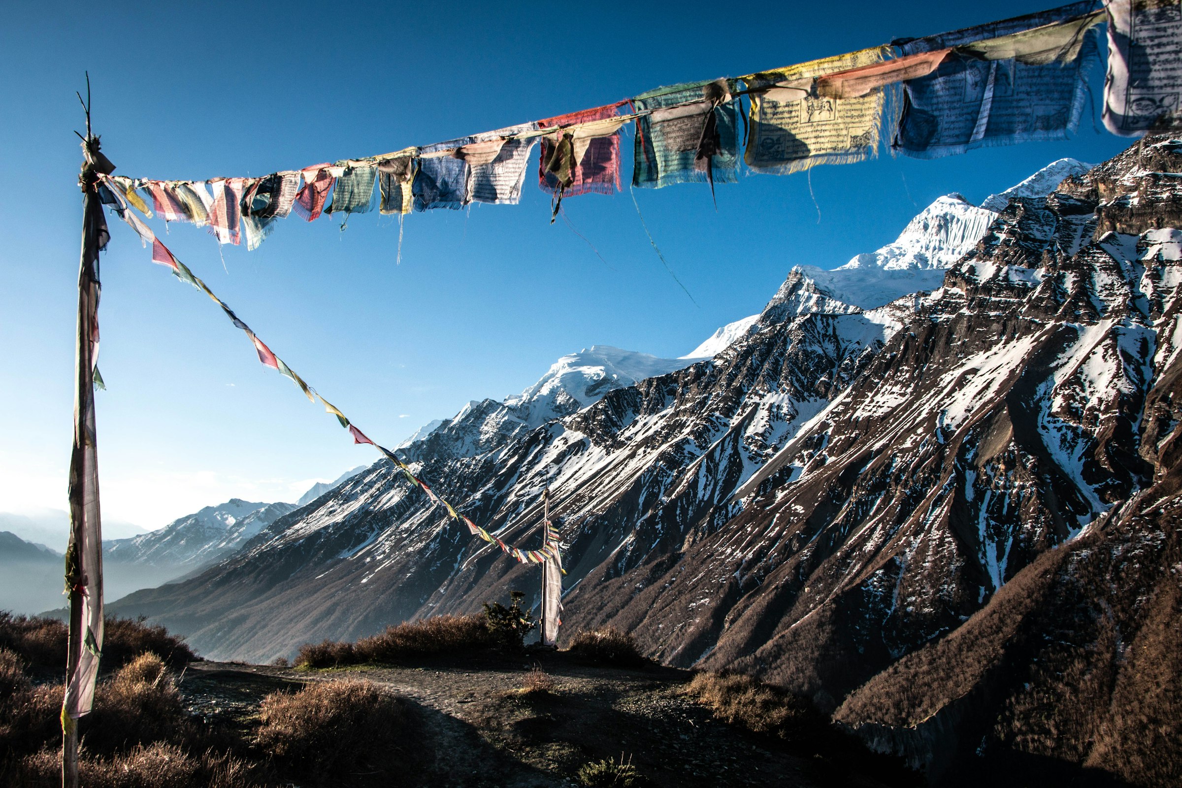 Prayer flags strung across a ridge above rugged snow-dusted mountains under a blue sky