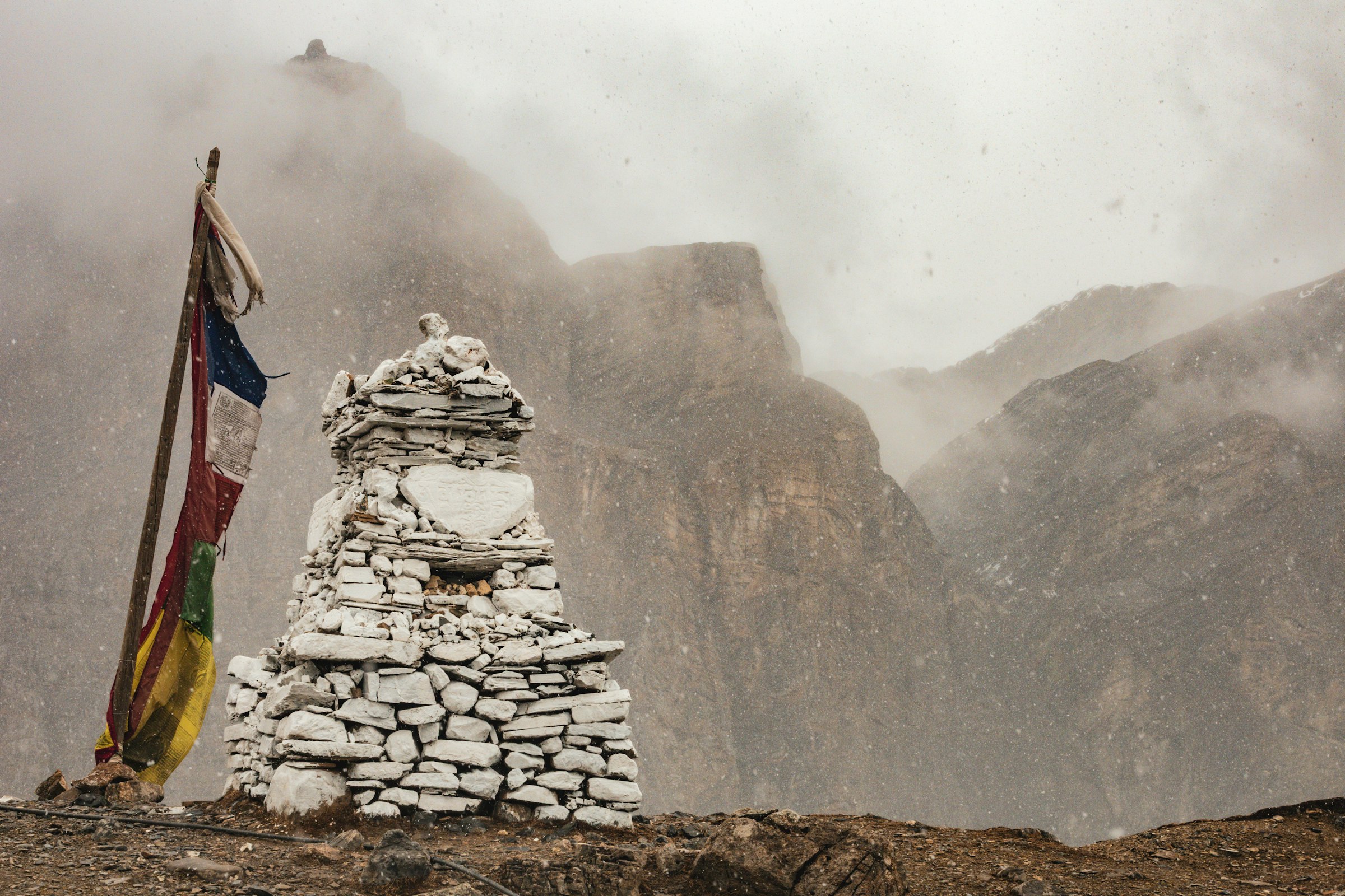 Stone chorten and prayer flag pole on a misty ridge before towering cliffs in the Annapurna region