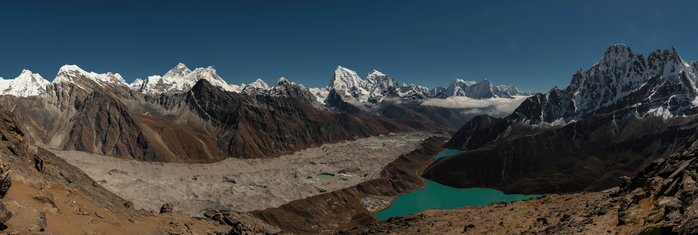 Wide panorama of the rubble-strewn Ngozumpa Glacier and turquoise Gokyo lake beneath snowy peaks