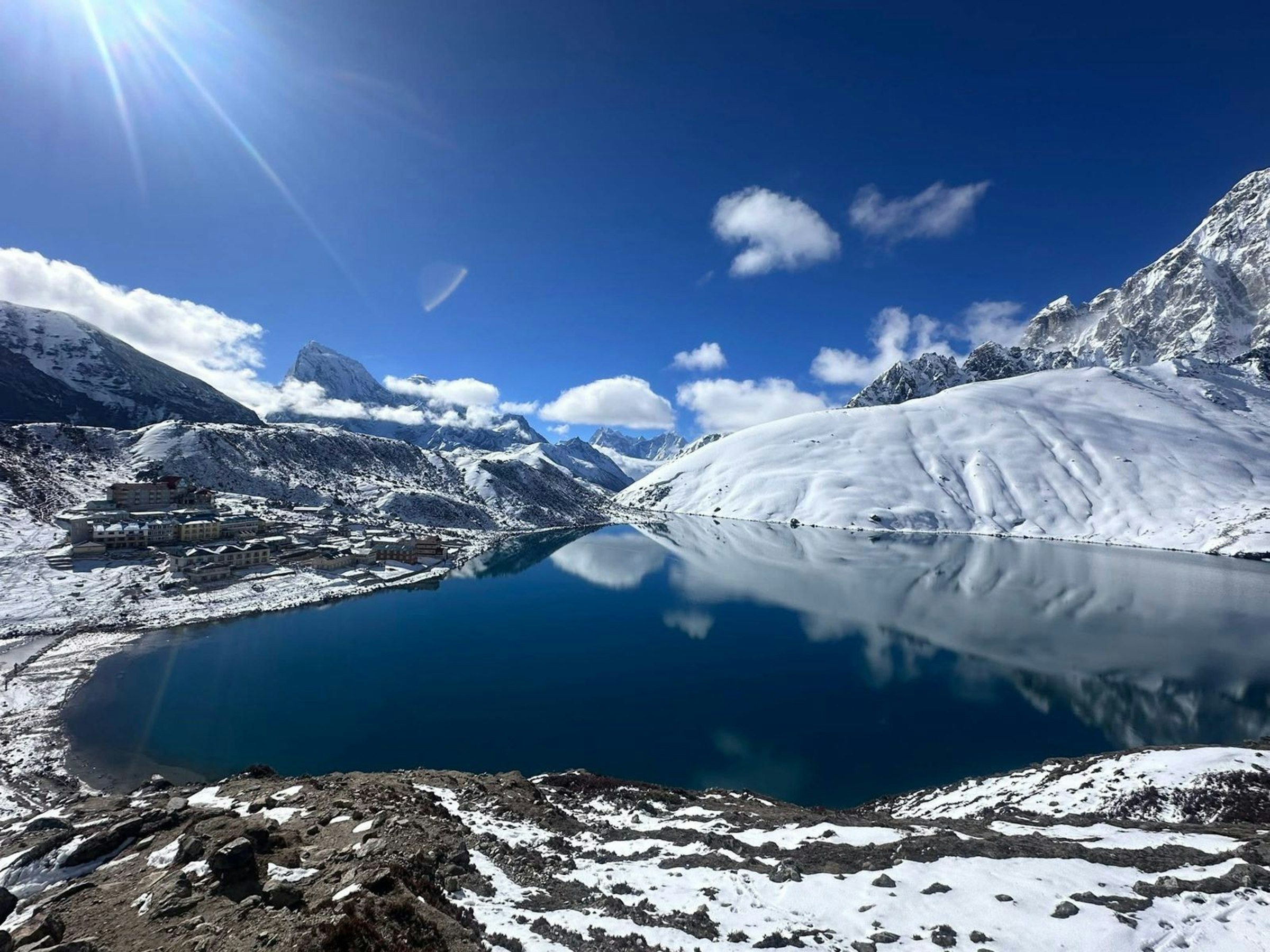Snowy slopes reflected in a deep blue lake under bright sunshine and scattered cloud in the Everest region