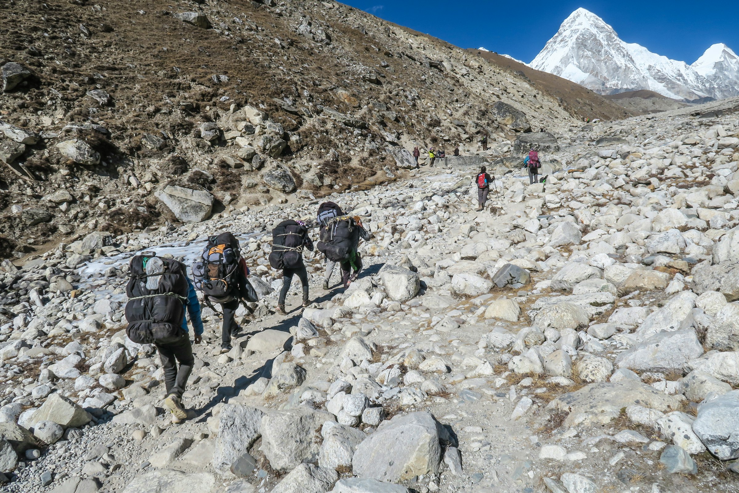 Porters with heavy loads walking a rocky moraine trail towards a snowy pyramid peak in the Khumbu