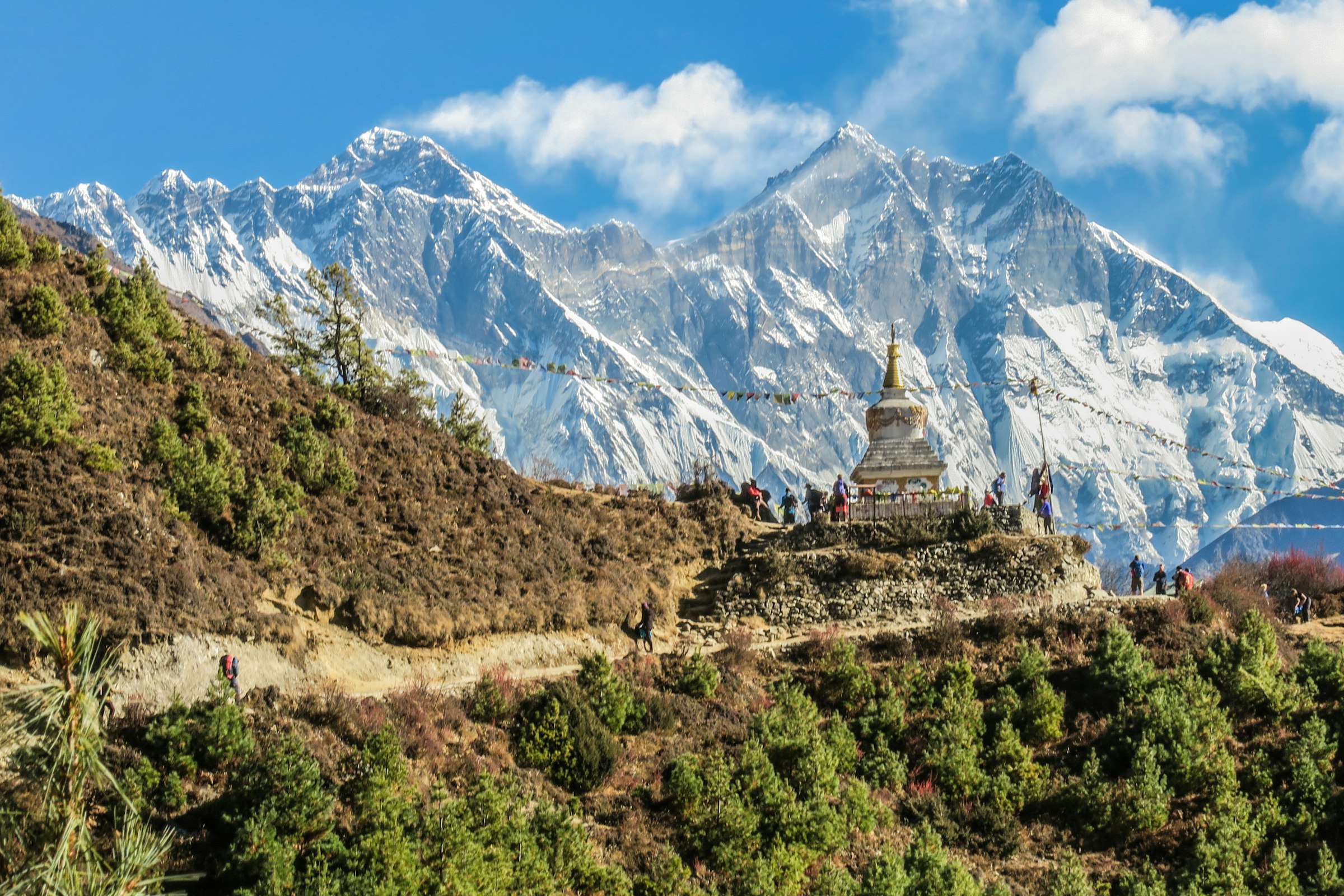 Trekkers gather at a stupa on a forested ridge with the Everest, Nuptse and Lhotse massif behind