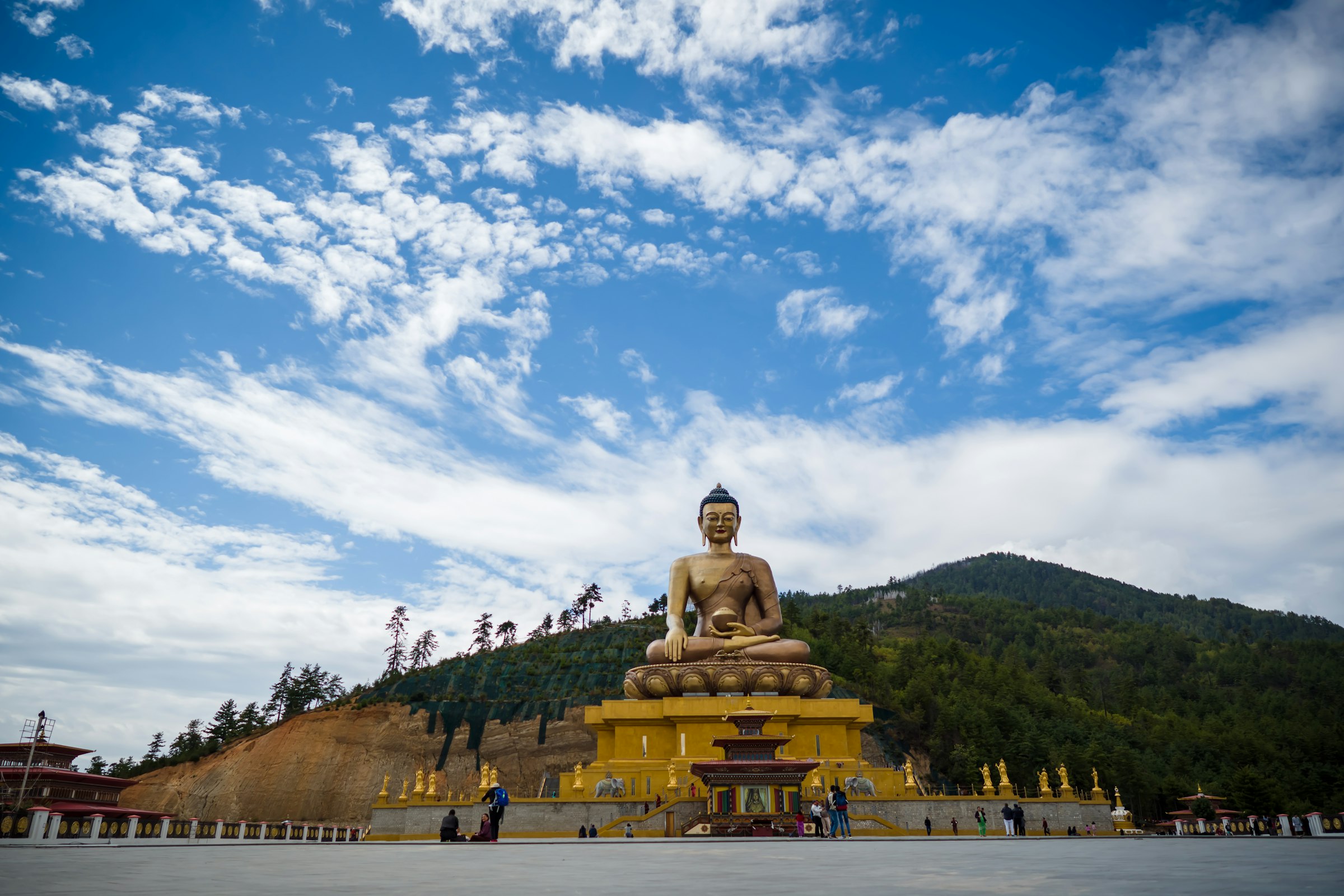 Giant golden Buddha Dordenma statue on a hillside platform under a cloud-streaked blue sky
