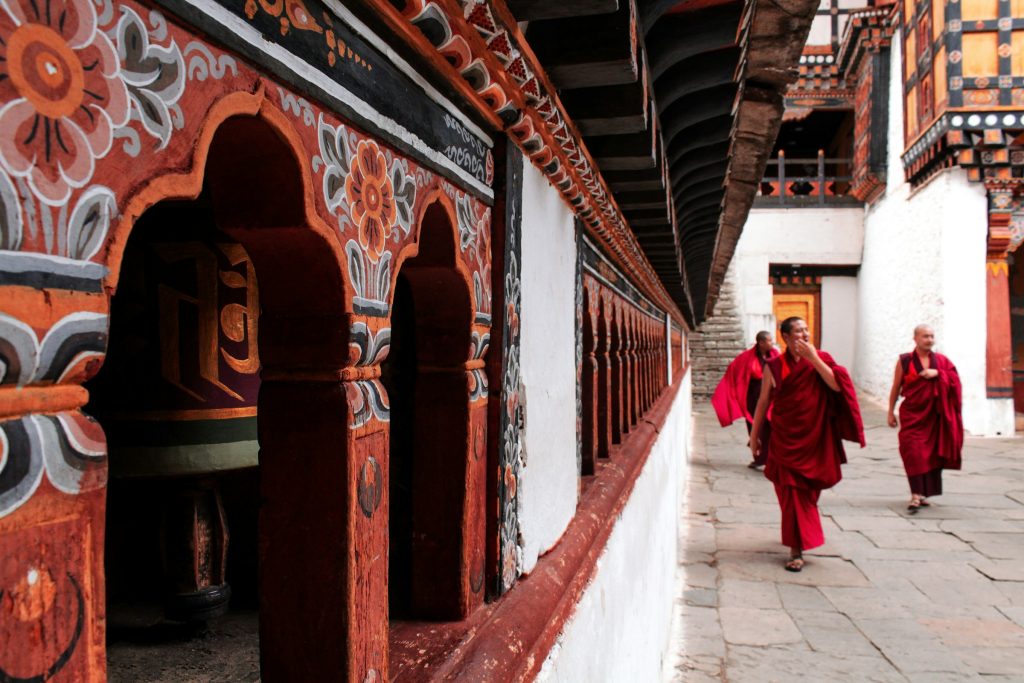 Two monks in maroon robes walking along a dzong corridor lined with painted wooden arches