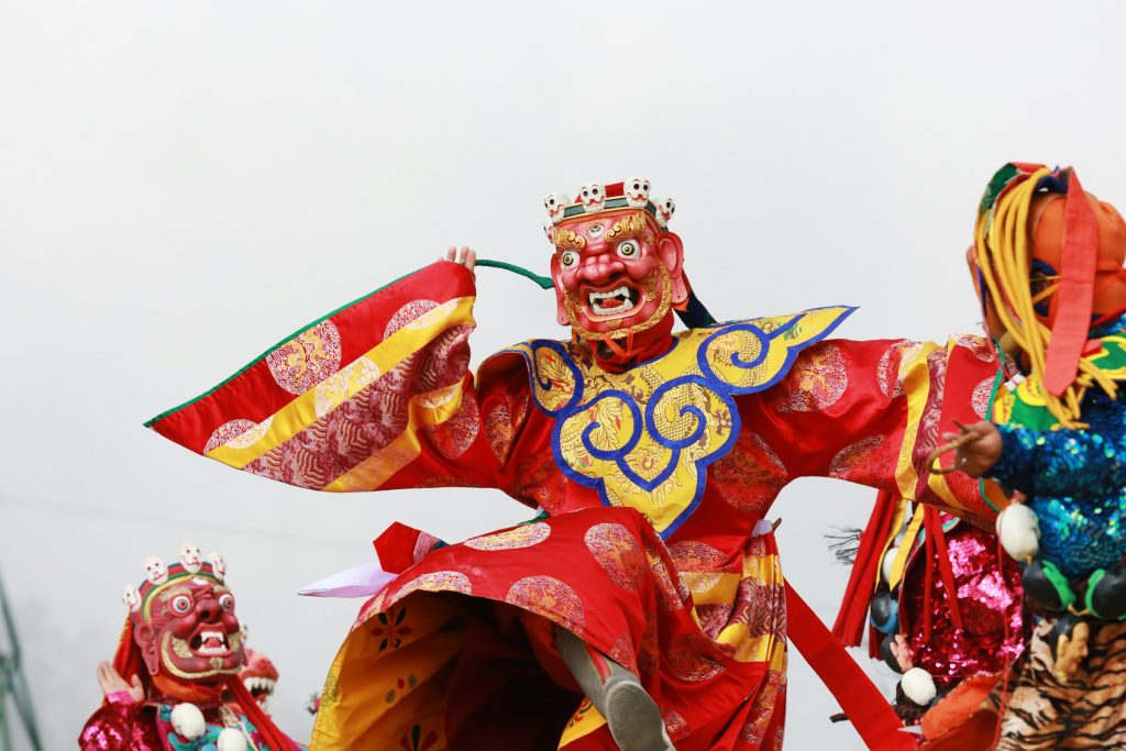 Masked dancer in a red and gold brocade costume performing a traditional festival dance