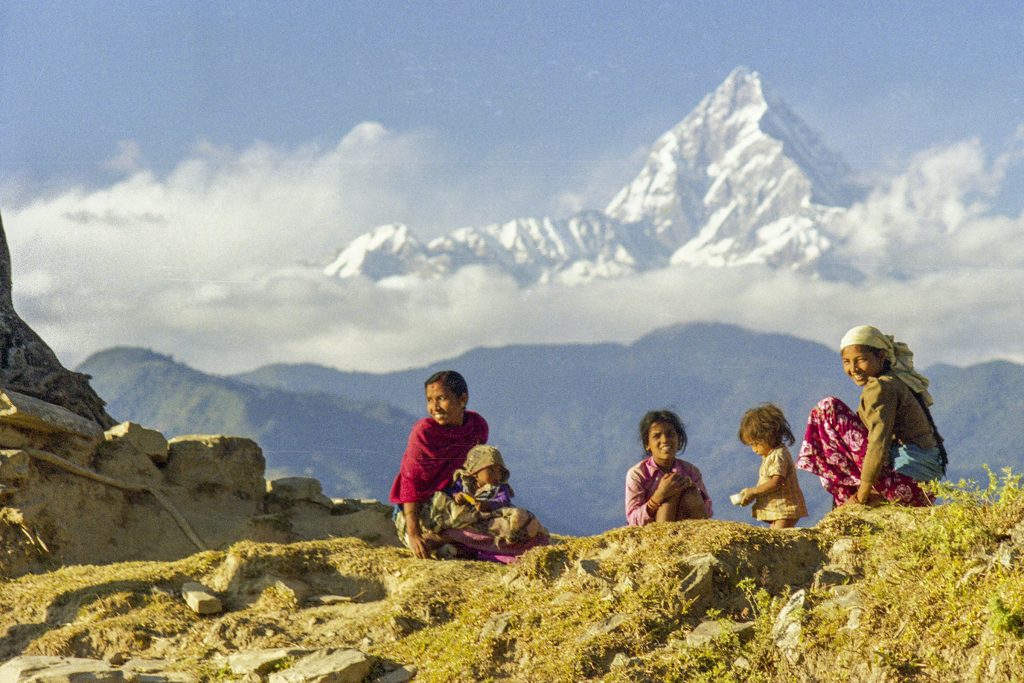 Village women and children sitting on a grassy hillside below Machhapuchhre's snowy summit