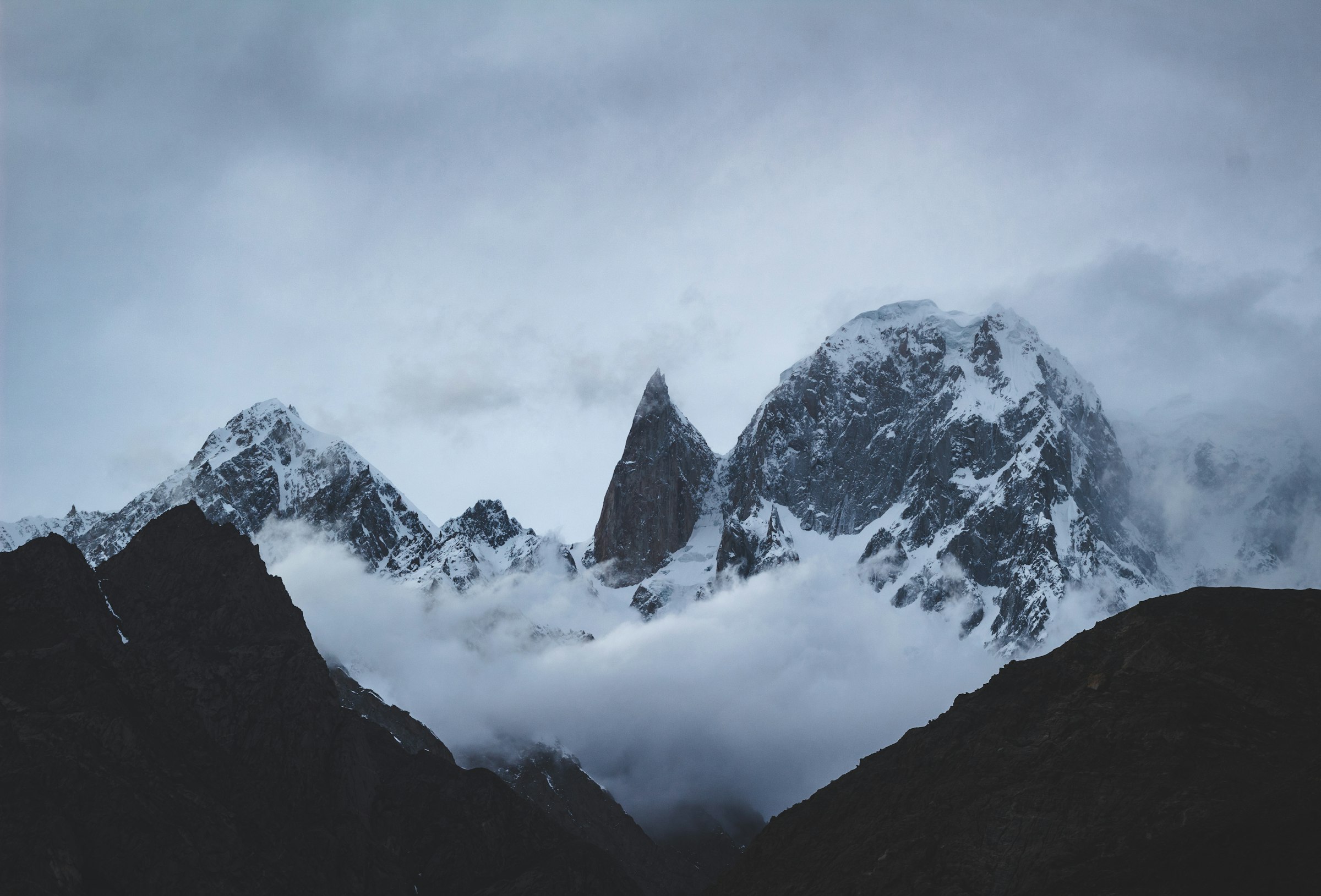 Pointed granite spire and snowy peaks emerging from swirling cloud above dark ridges in the Karakoram