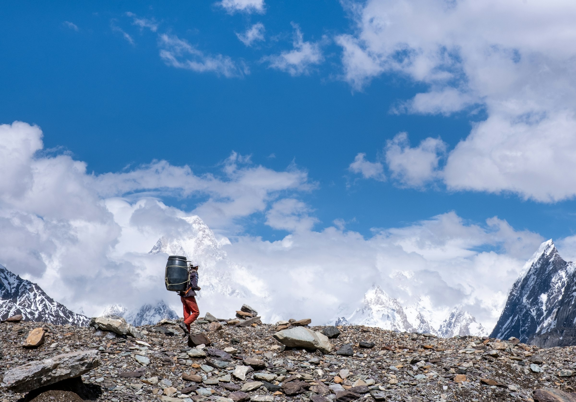 Porter carrying a blue barrel across a rocky glacier moraine beneath billowing clouds in the Karakoram