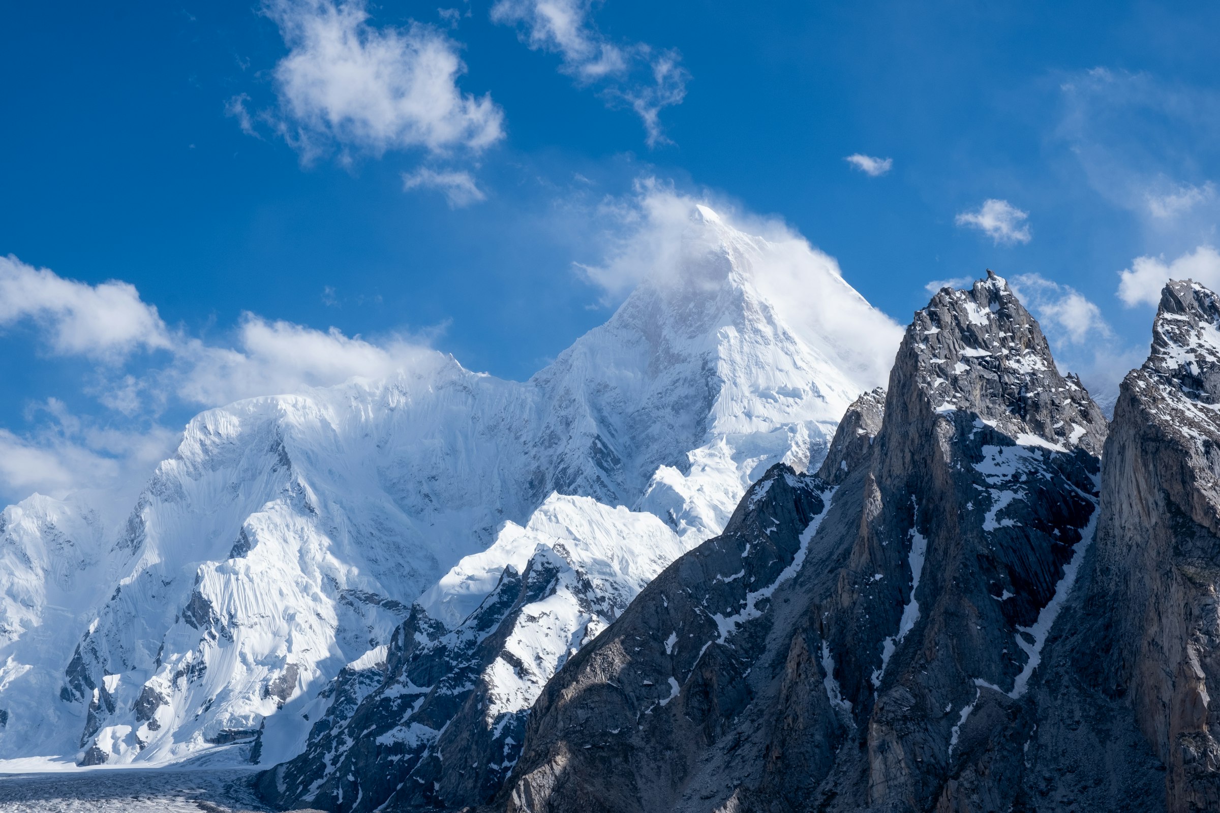 Cloud-capped snowy peak rising behind dark granite spires under a blue sky in the Karakoram