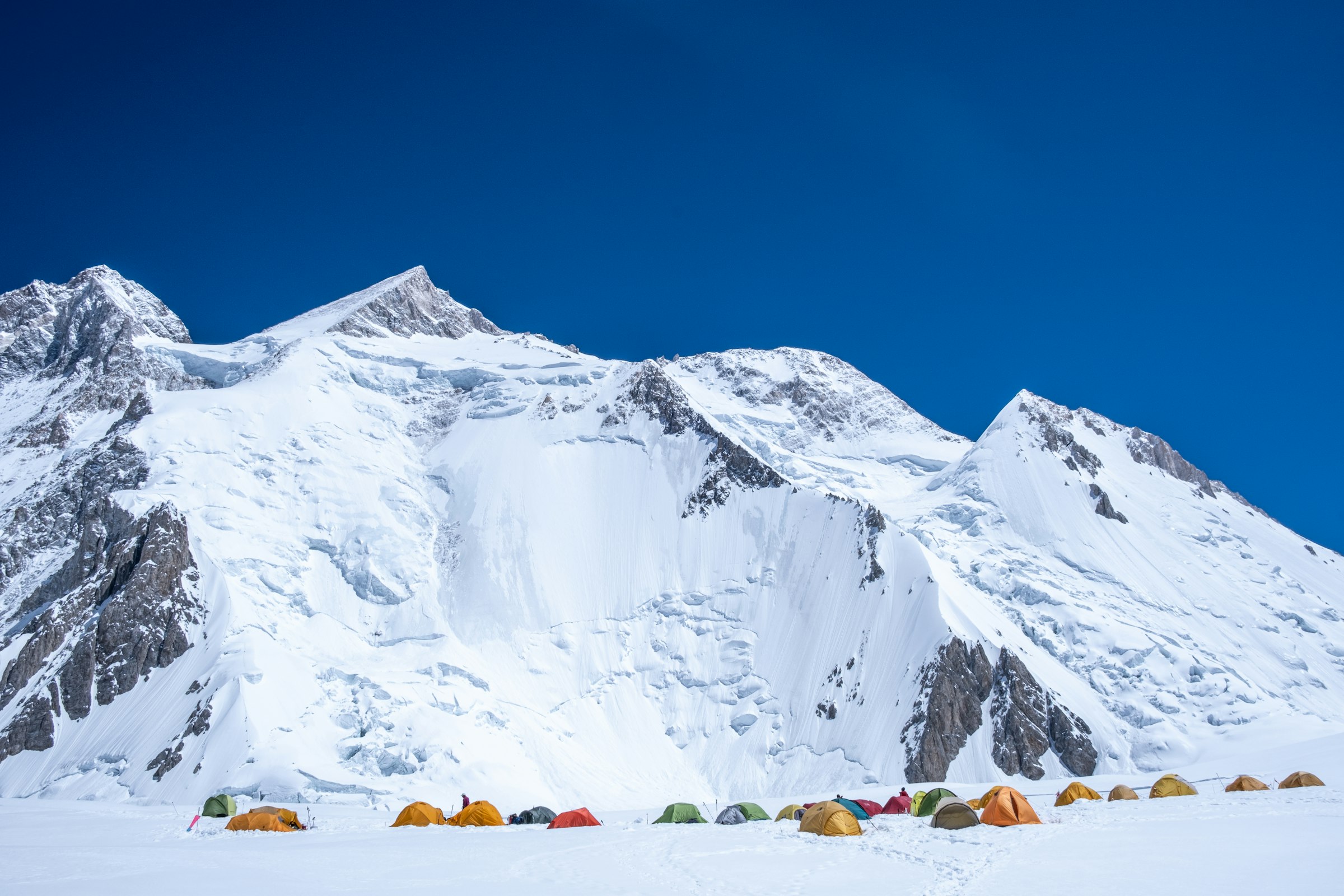 Colourful expedition tents on a snowy glacier below a huge white mountain under a deep blue sky