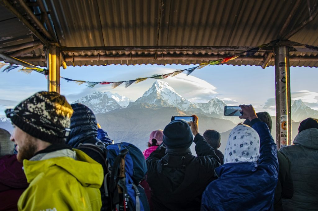 Trekkers in warm hats photographing snowy peaks at sunrise from a covered viewpoint strung with prayer flags
