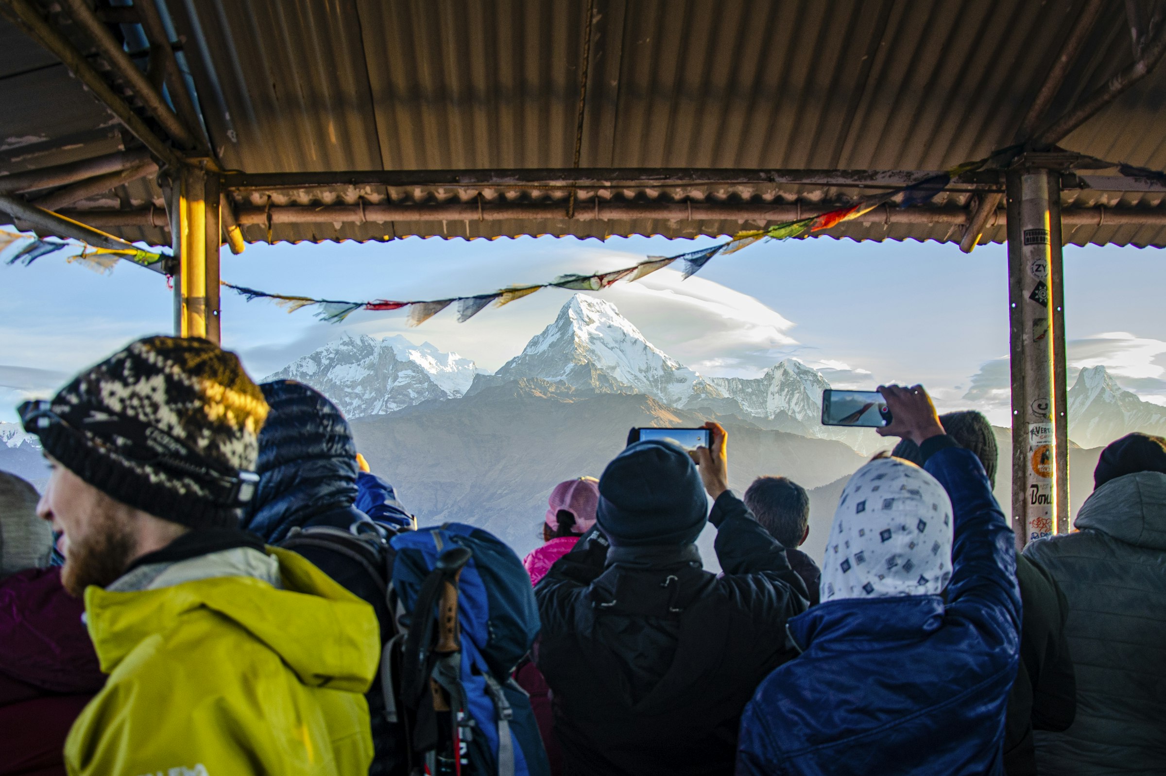 Trekkers in warm hats photographing snowy peaks at sunrise from a covered viewpoint strung with prayer flags