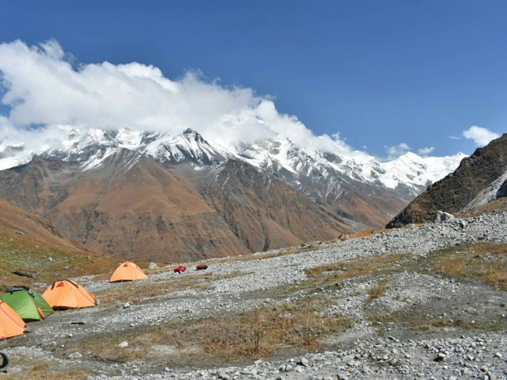 Orange and green tents pitched on a stony plain below brown hills and a snowy range