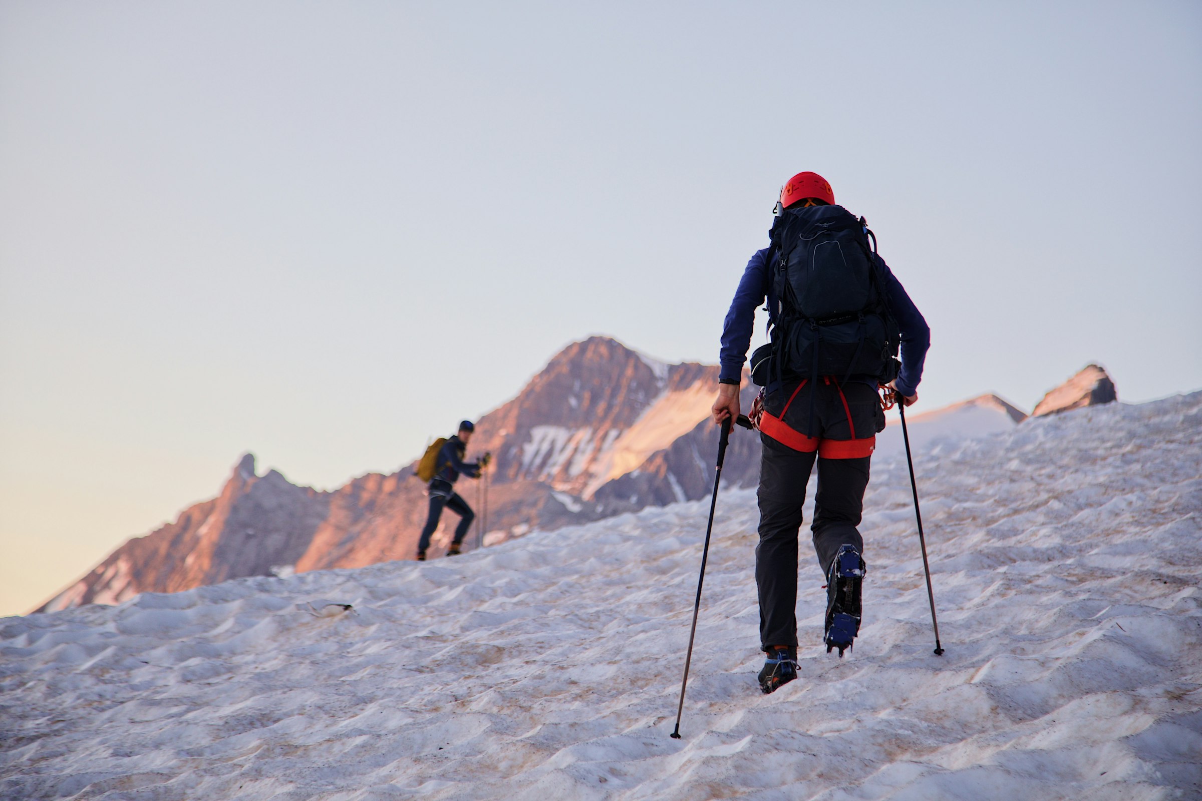 Two climbers with trekking poles ascending a snow slope towards rocky peaks lit pink at dawn