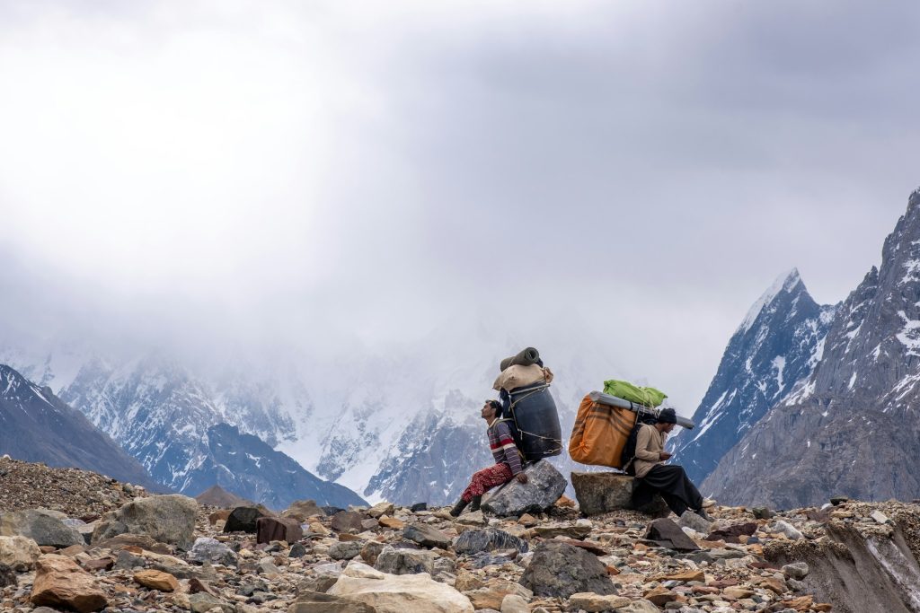 Porters resting with their loads on a rocky moraine below cloud-wrapped jagged peaks in the Karakoram