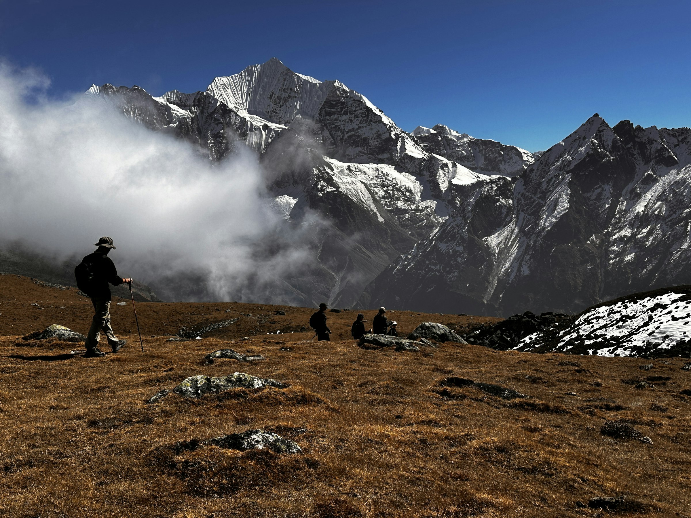 Trekkers crossing a brown alpine meadow below a snow-capped peak and drifting cloud in the Langtang region
