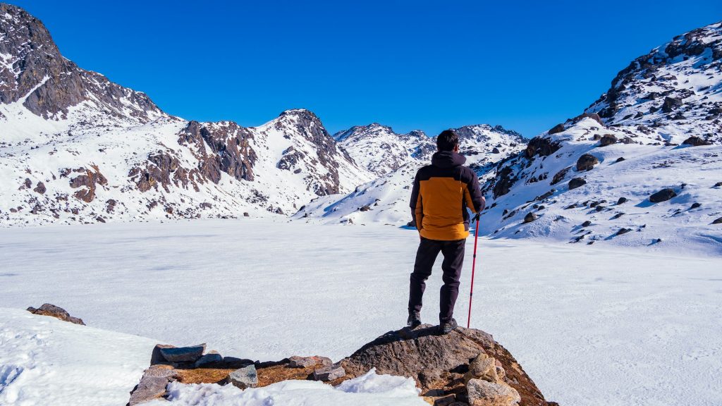 Trekker in an orange jacket standing on a rock above a snow-covered frozen lake ringed by peaks