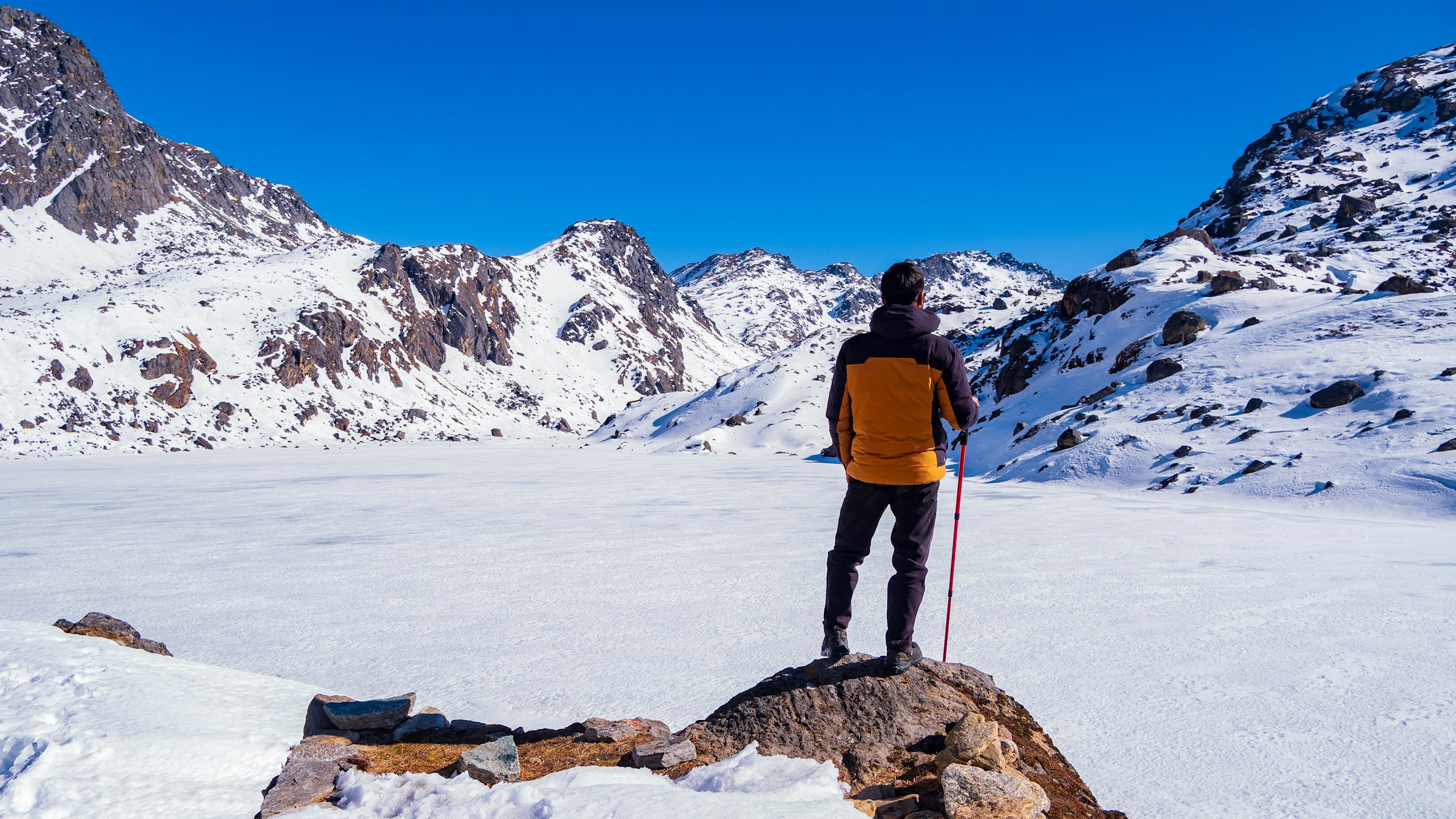 Trekker in an orange jacket standing on a rock above a snow-covered frozen lake ringed by peaks
