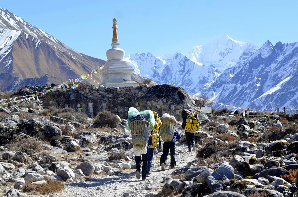 Porters carrying heavy loads along a rocky trail towards a white stupa below snowy peaks in Langtang