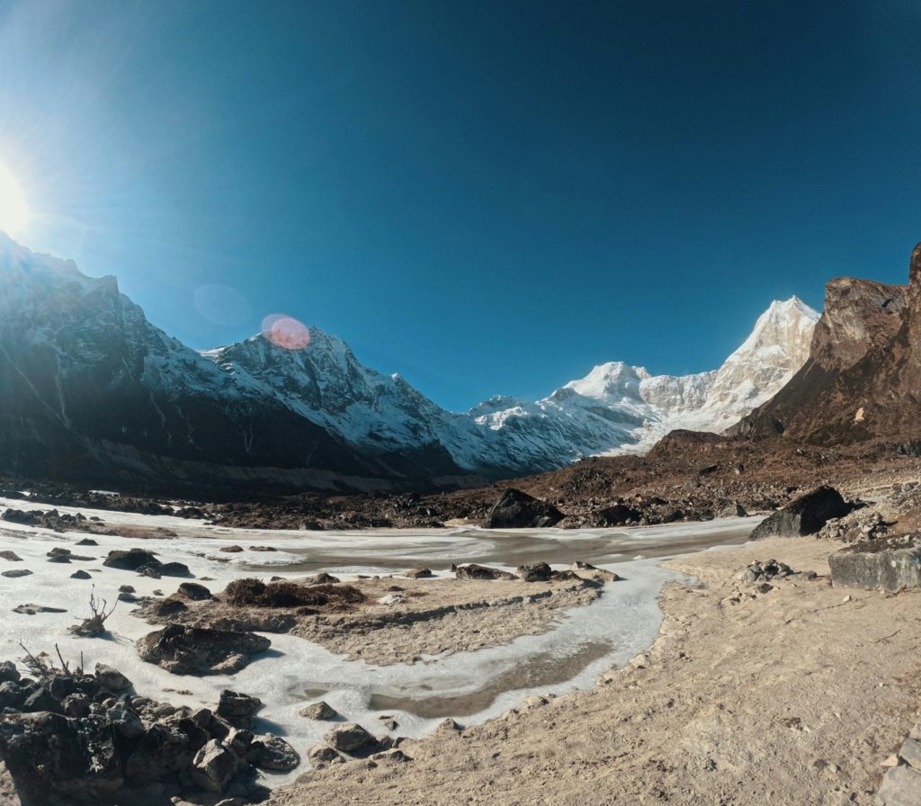 Frozen stream crossing a sunlit valley floor below snowy peaks in the Manaslu region