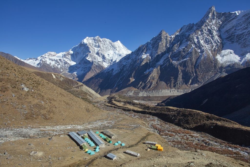 Cluster of tin-roofed lodges on a brown hillside below jagged snowy peaks in the Manaslu region