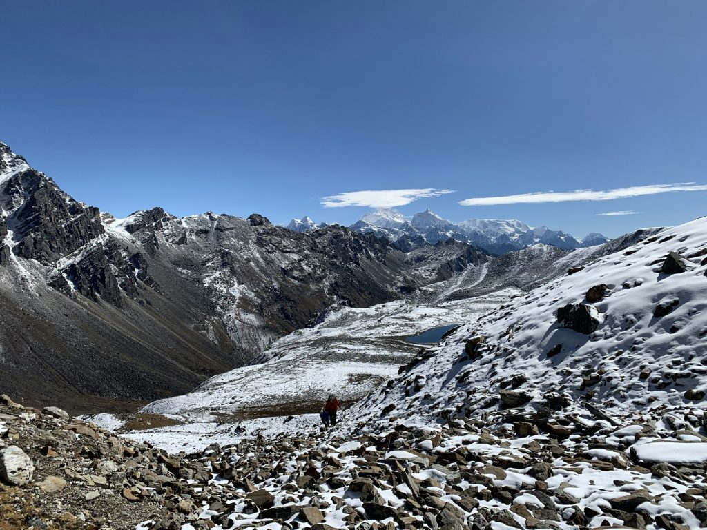 Lone trekker crossing a rocky, snow-dusted slope above a high valley with distant peaks