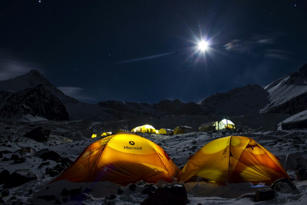 Glowing yellow dome tents at a rocky high camp under a bright moon and starry sky