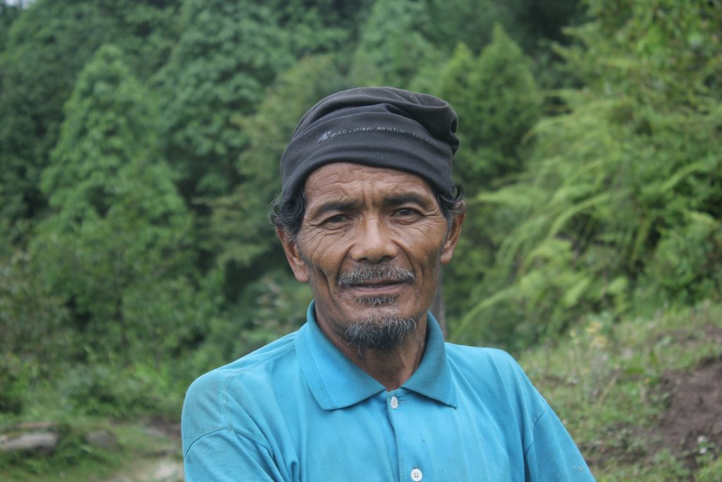 Portrait of a weathered local man in a dark headscarf and turquoise shirt beside green forest