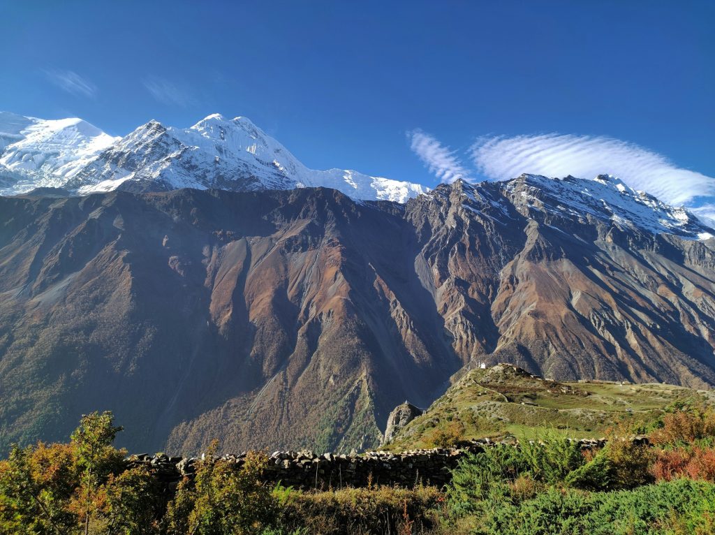 Snow-capped peaks above vast brown slopes with a stone wall and shrubs in the foreground
