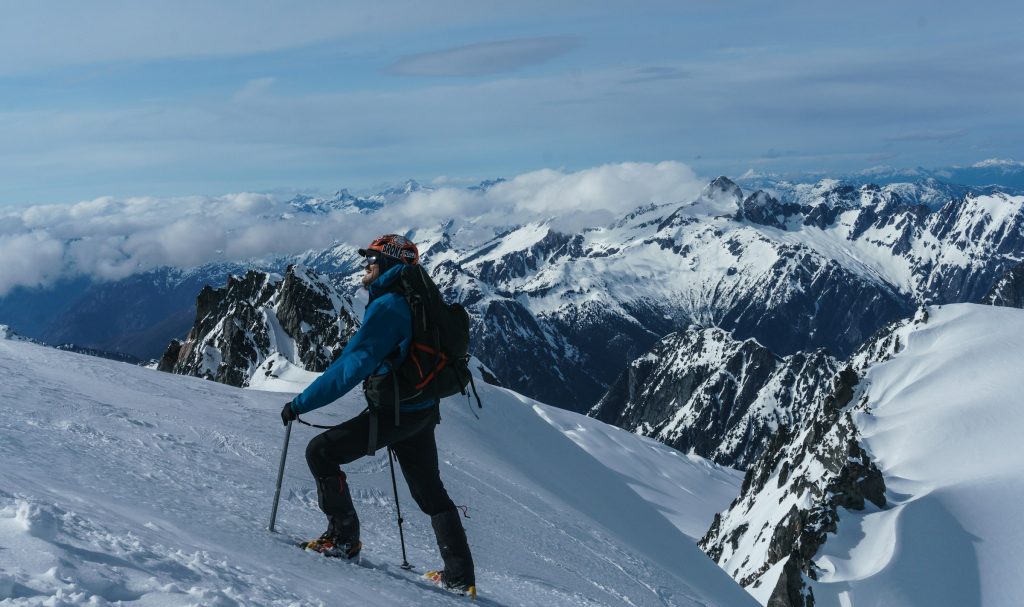 Climber in a blue jacket with trekking poles on a snowy ridge above a sea of peaks