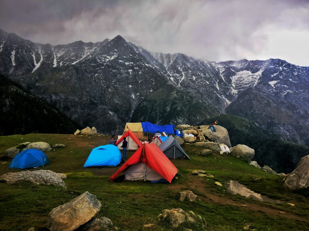 Colourful tents pitched on a grassy meadow among boulders below dark snow-streaked mountains