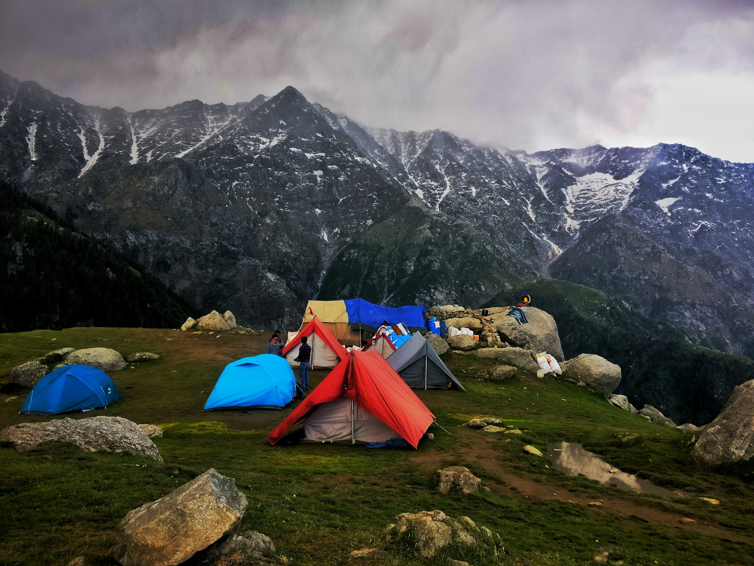 Colourful tents pitched on a grassy meadow among boulders below dark snow-streaked mountains