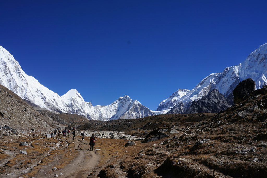Line of trekkers walking a dusty trail through a rocky valley towards snowy peaks
