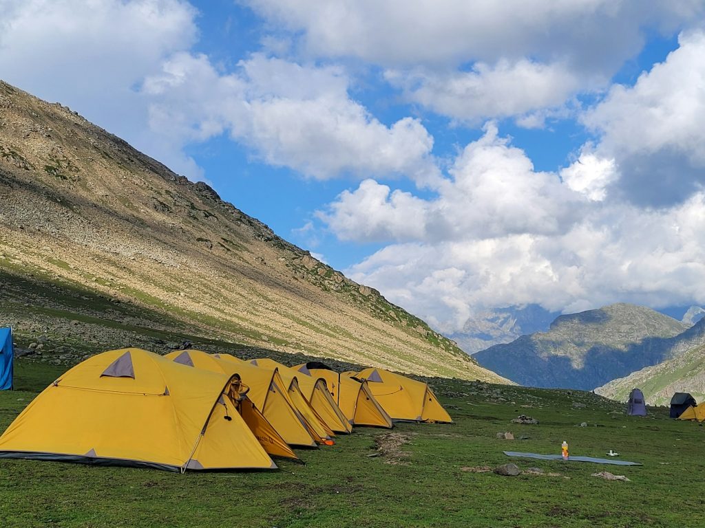 Row of yellow dome tents pitched on a green meadow beneath grassy slopes and white clouds