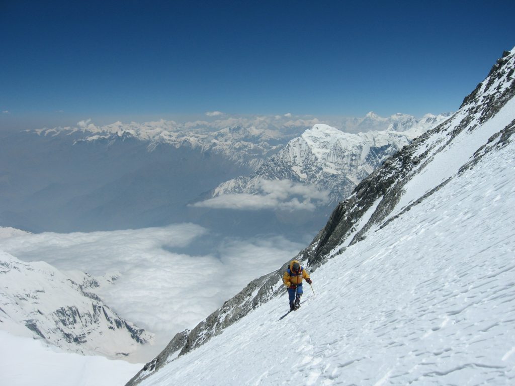 Climber in a yellow jacket ascending a steep snow slope high above a sea of cloud
